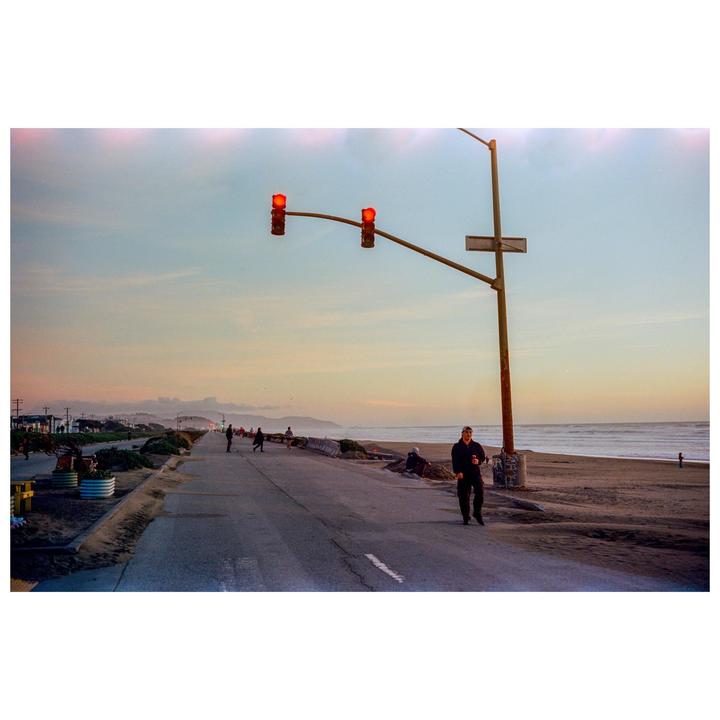 A man walks along a coastal road with a beach and ocean to his right, and a traffic light overhead.