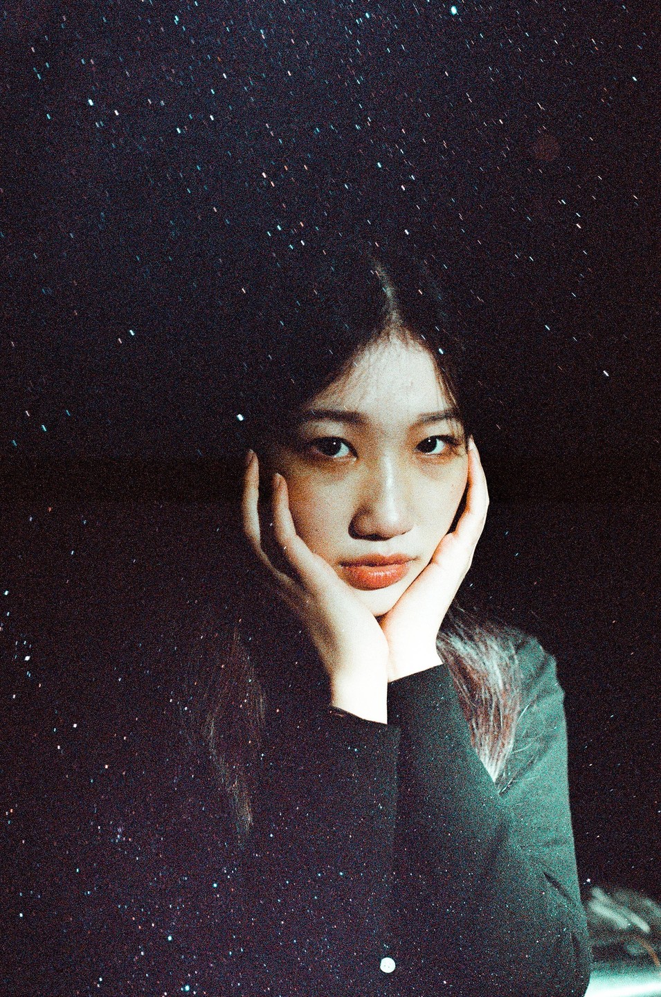 A woman with long dark hair rests her chin on her hands, looking directly at the viewer against a dark, star-filled background.