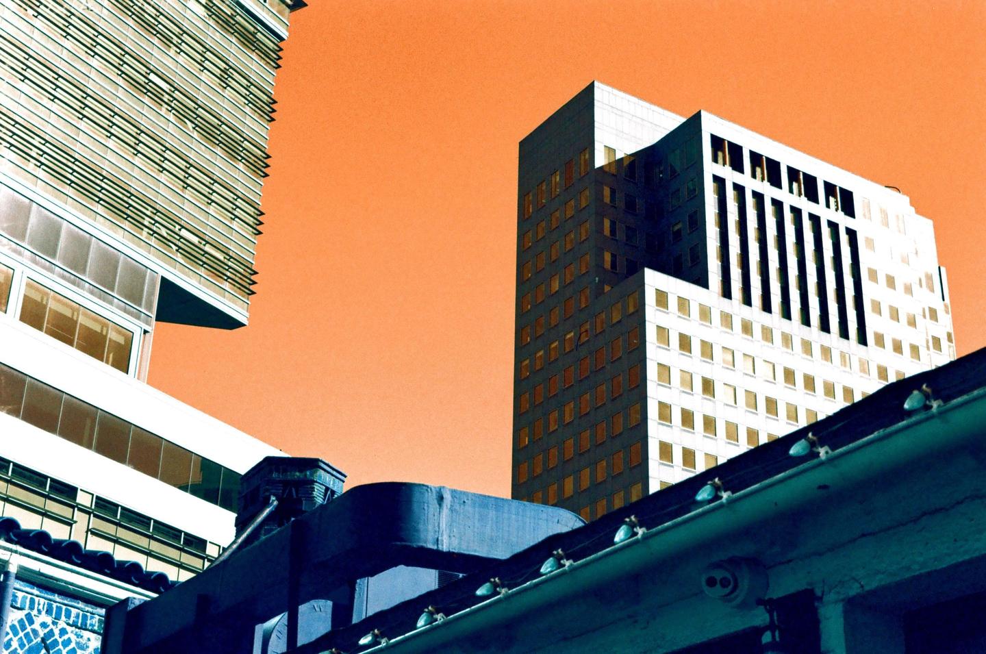 Two buildings with many windows are visible against an orange sky, with a dark blue structure in the foreground.