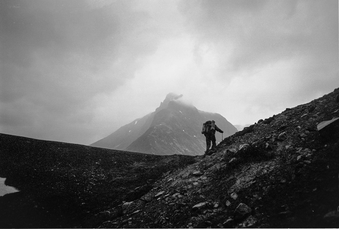 A hiker with a backpack and walking stick ascends a rocky mountain path under a cloudy sky.