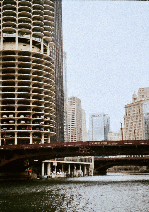 A tall, cylindrical building with visible parking spaces on each floor stands next to a river with a bridge crossing it, with other buildings in the background.