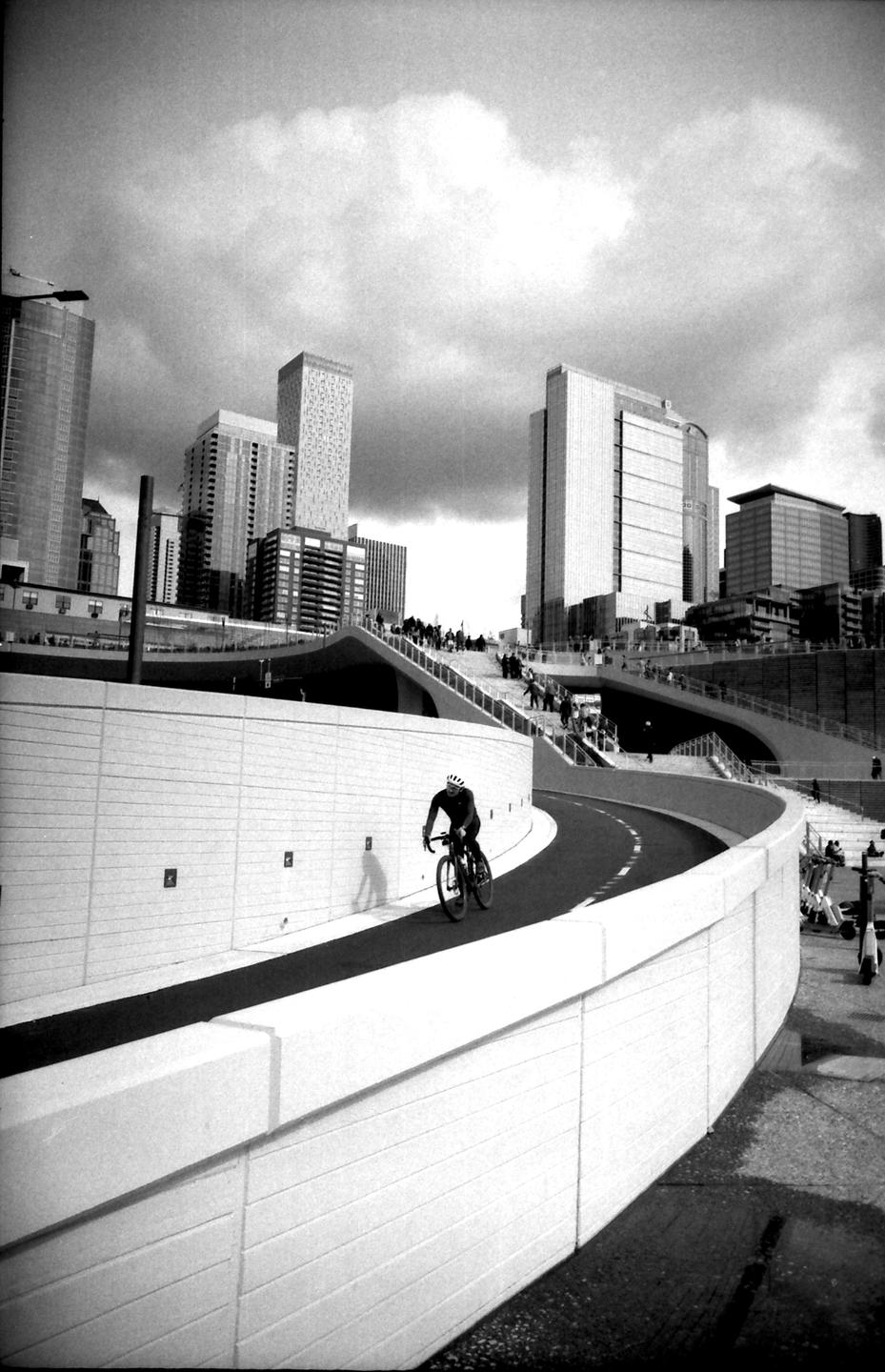 A cyclist rides on a curved path next to a wall, with city buildings and people on stairs in the background.