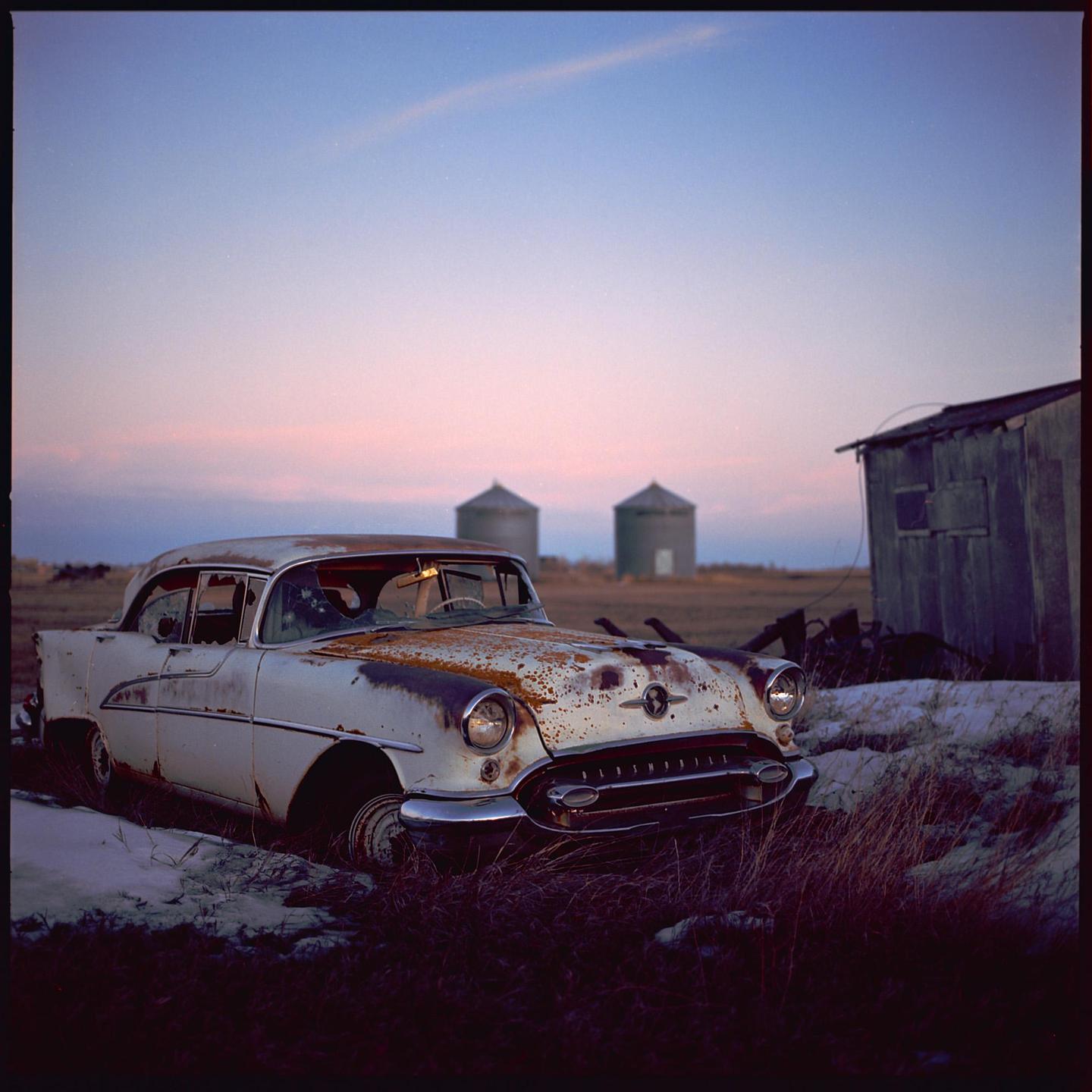 A rusty white car is parked in a field with two silos and a small shed under a colorful sky.