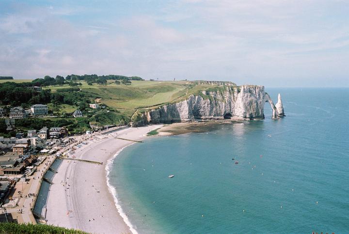 A wide view shows a beach town nestled against a cliff with a natural arch formation extending into the sea.