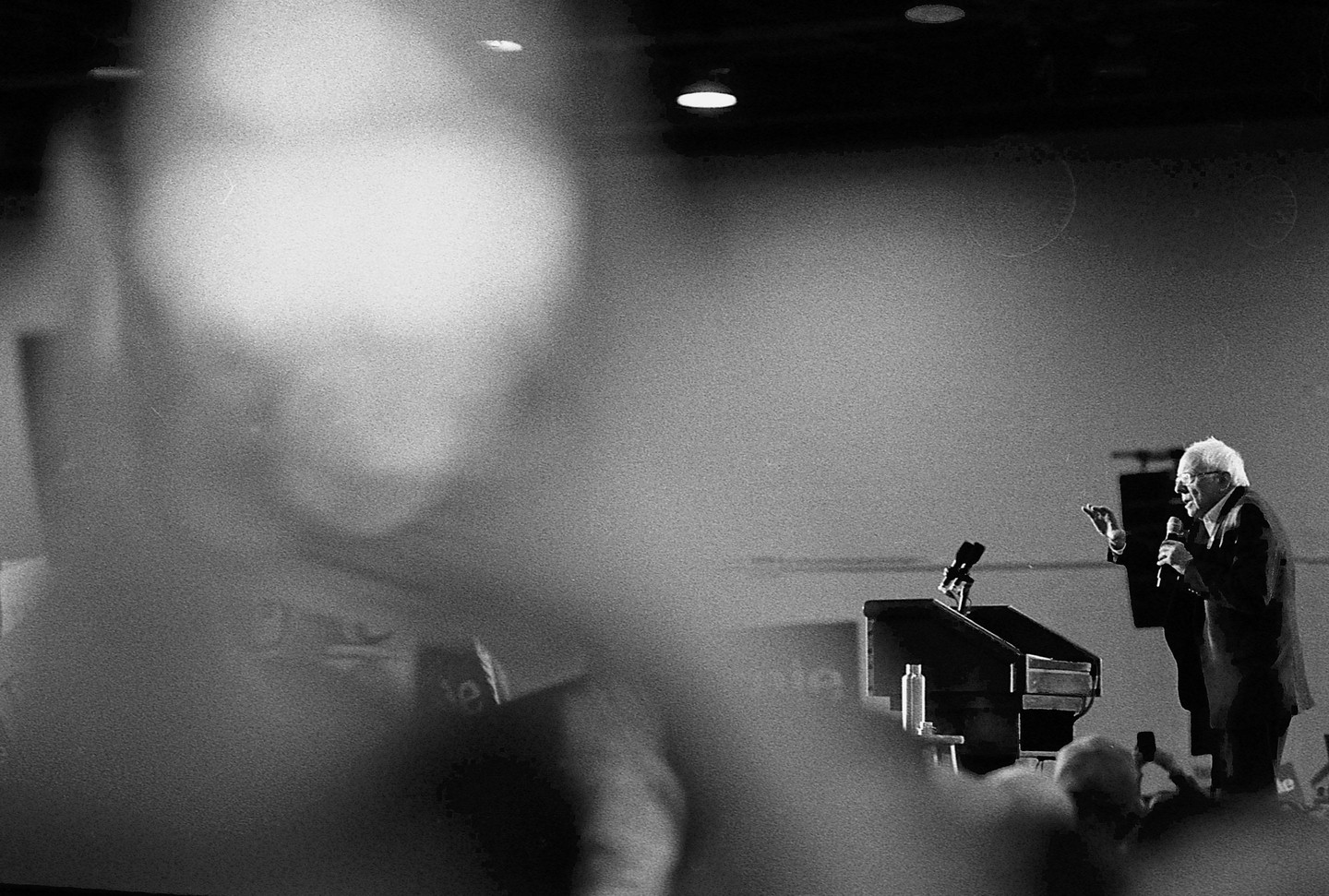 A man speaks into a microphone at a podium on a stage, with a blurred audience in the foreground.