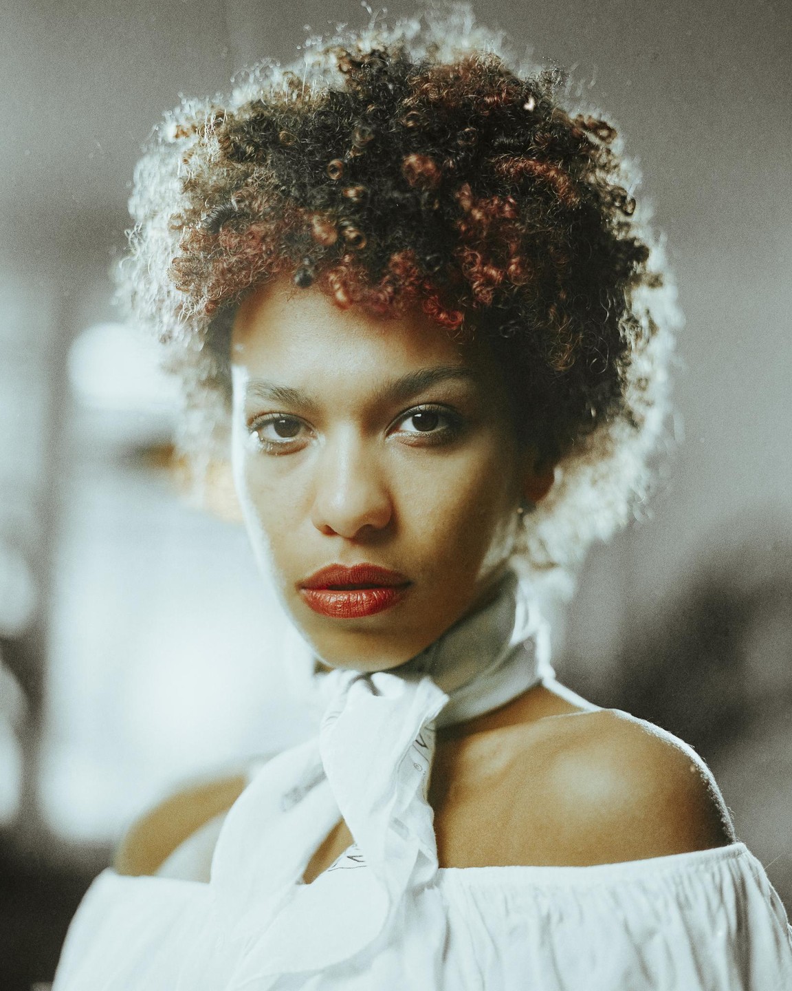 A woman with curly hair and red lipstick wears a white off-the-shoulder top and a white scarf.