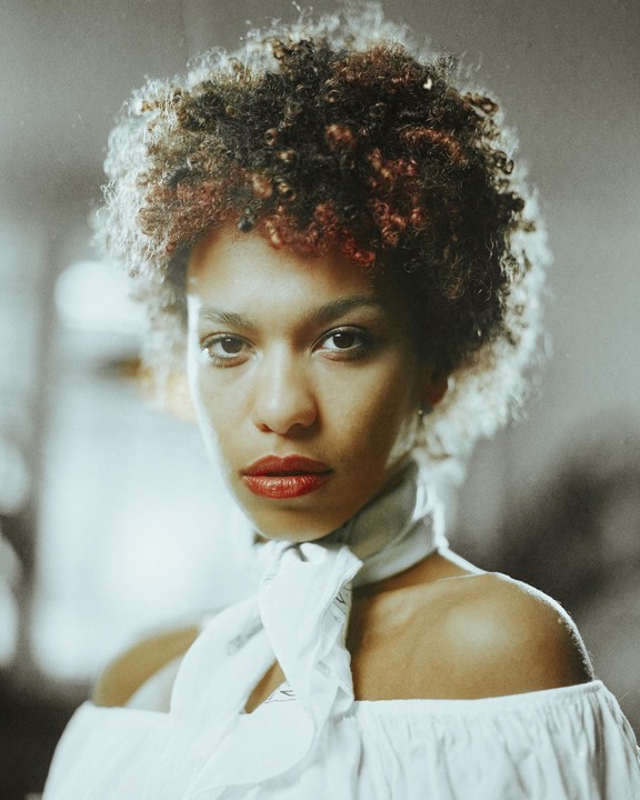 A woman with curly hair and red lipstick wears a white off-the-shoulder top and a white scarf.