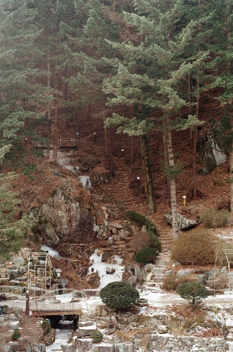 A winding path with stairs ascends a snowy, forested hill with a frozen waterfall and a water wheel.
