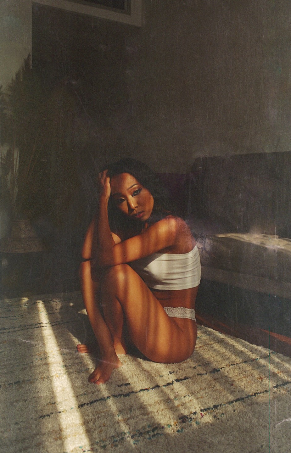A woman sits on a rug with her knees pulled to her chest, looking at the viewer as light streams across her body.
