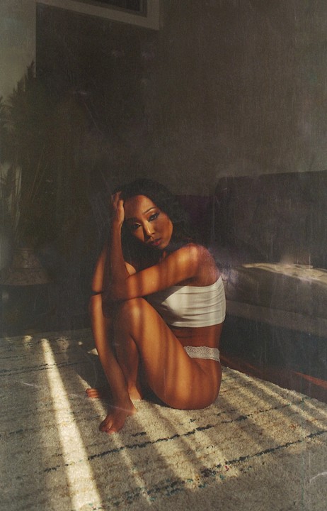 A woman sits on a rug with her knees pulled to her chest, looking at the viewer as light streams across her body.