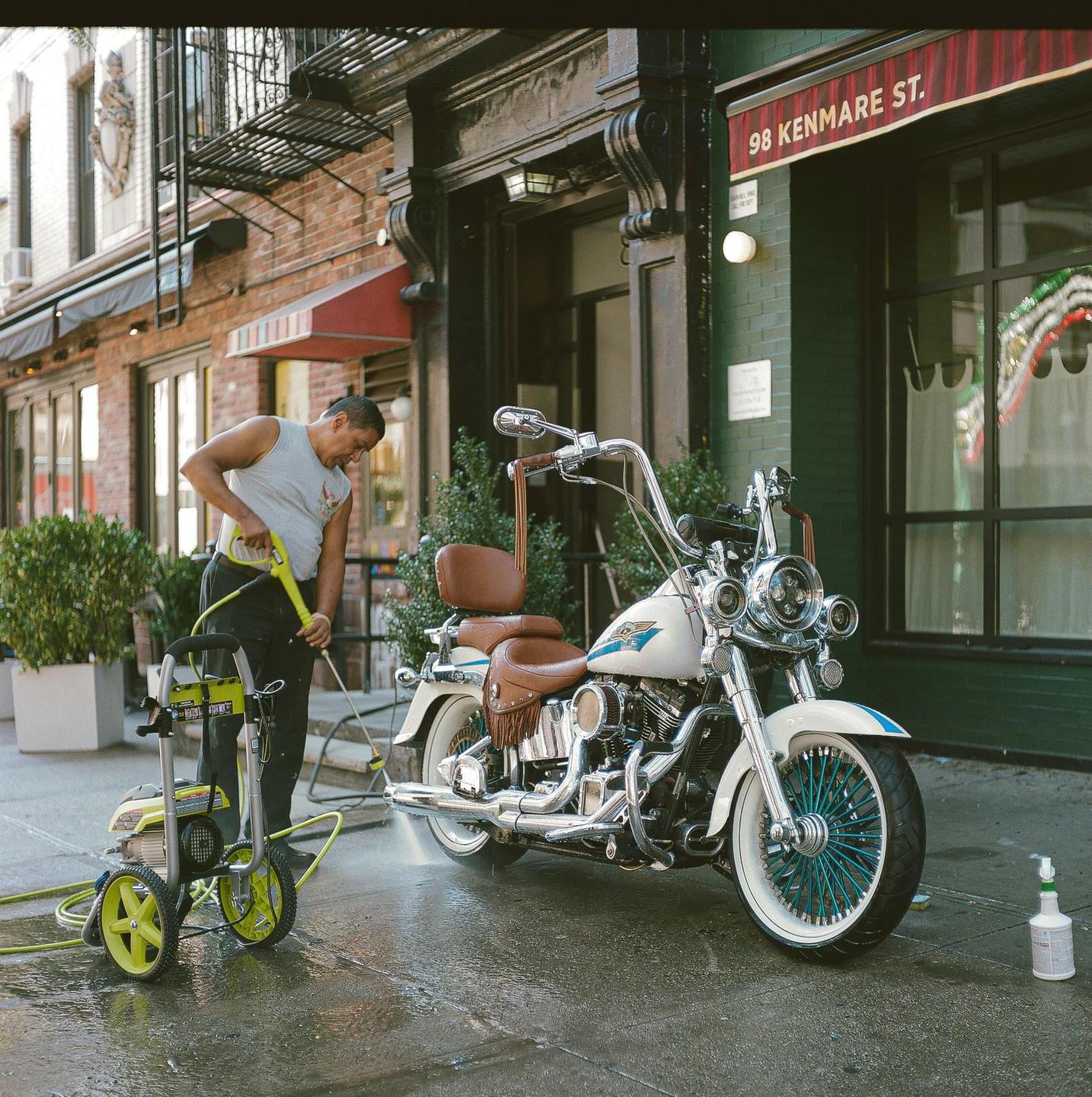 A man washes a white motorcycle with a pressure washer on a city street.