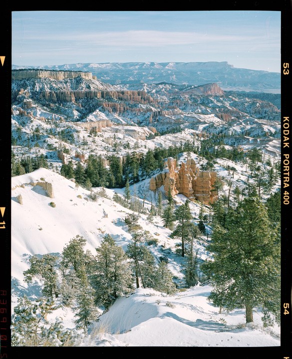 A vast snowy landscape features a canyon with rock formations and evergreen trees under a clear sky.
