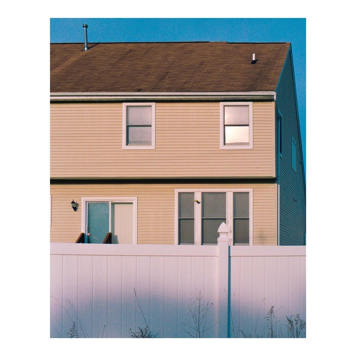 A beige house with a brown roof is visible behind a white fence under a clear blue sky.