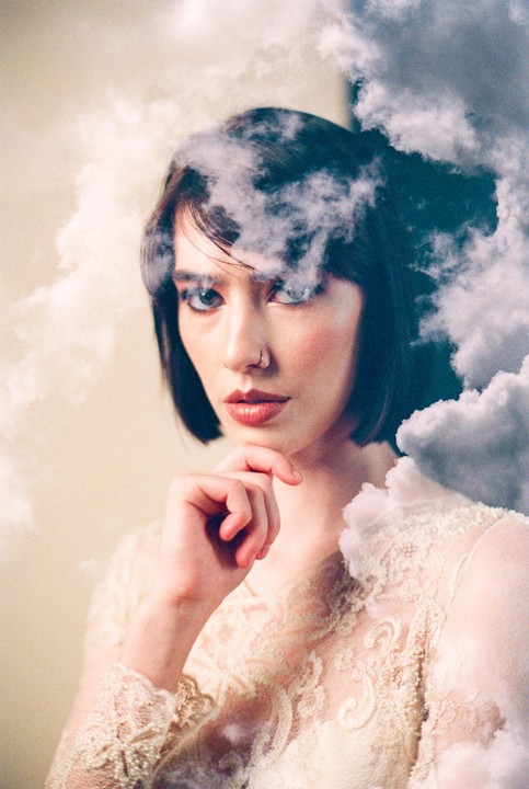 A woman with dark hair and a nose ring looks directly at the viewer, surrounded by clouds.