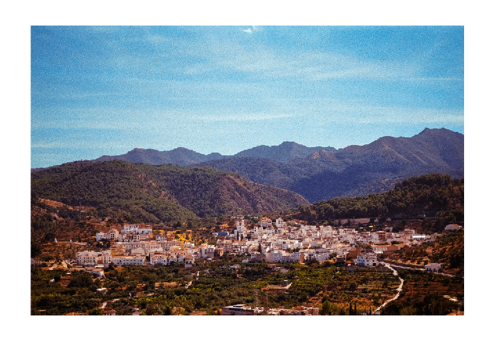 A village with white buildings is nestled in a valley surrounded by mountains and green hills under a clear blue sky.