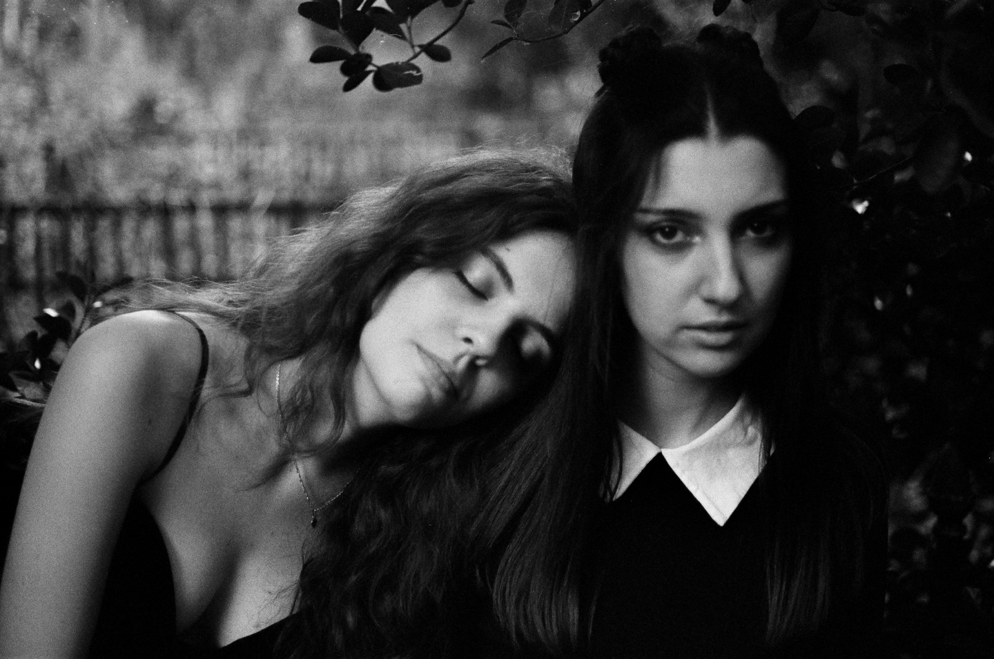 One woman rests her head on another woman's shoulder, both looking at the viewer.