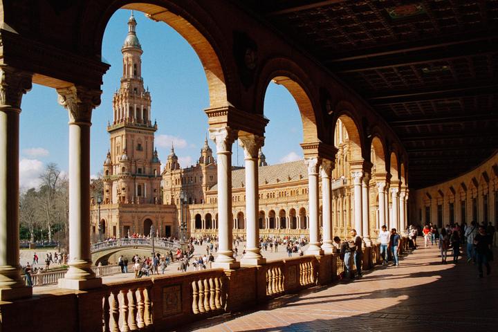 A large, ornate building with a prominent tower is visible through a series of arches, with many people walking around a plaza in front of it.