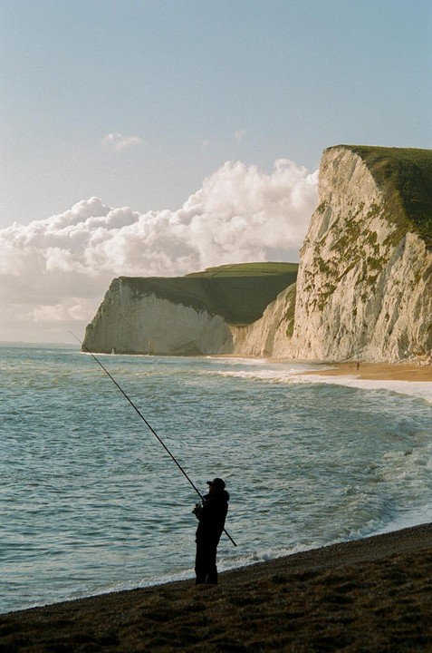 A fisherman stands on a rocky beach with a fishing rod, facing the ocean with cliffs in the background.