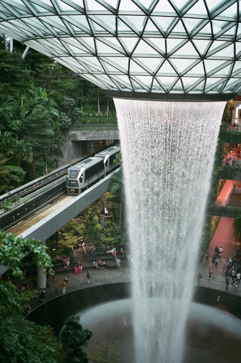 A train passes by a large indoor waterfall under a glass dome, surrounded by lush greenery and people.