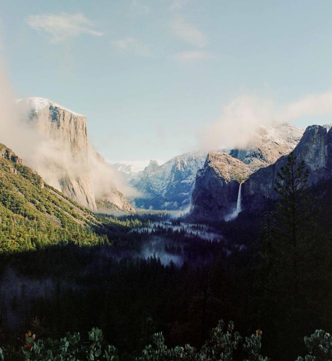 A valley with a waterfall and a forest is surrounded by mountains partially covered in clouds.