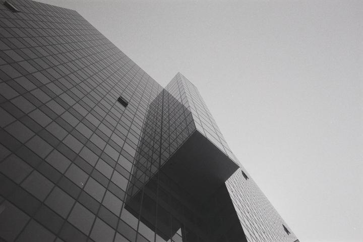A low-angle view shows the glass facade of a modern building reflecting another building against a bright sky.