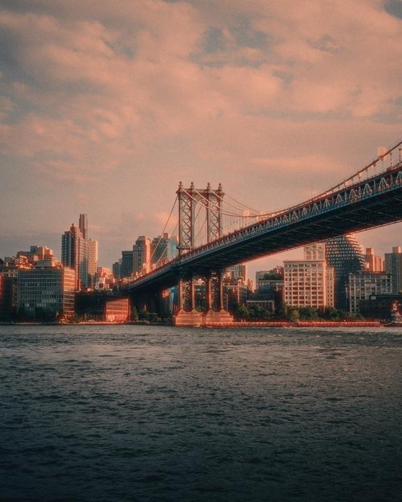 The Manhattan Bridge spans a body of water with a city skyline in the background under a cloudy sky.