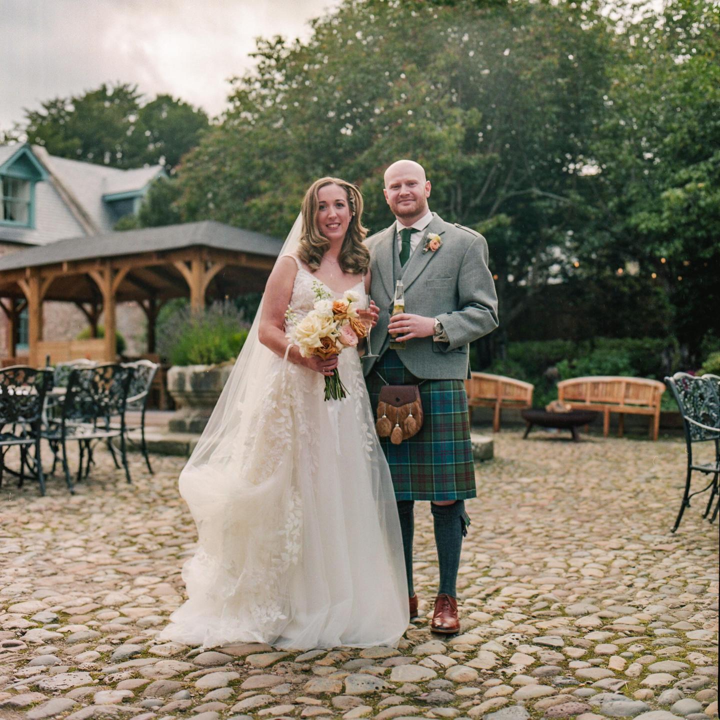 A couple stands on a cobblestone patio, the woman in a wedding dress holding a bouquet and the man in a kilt holding a bottle.