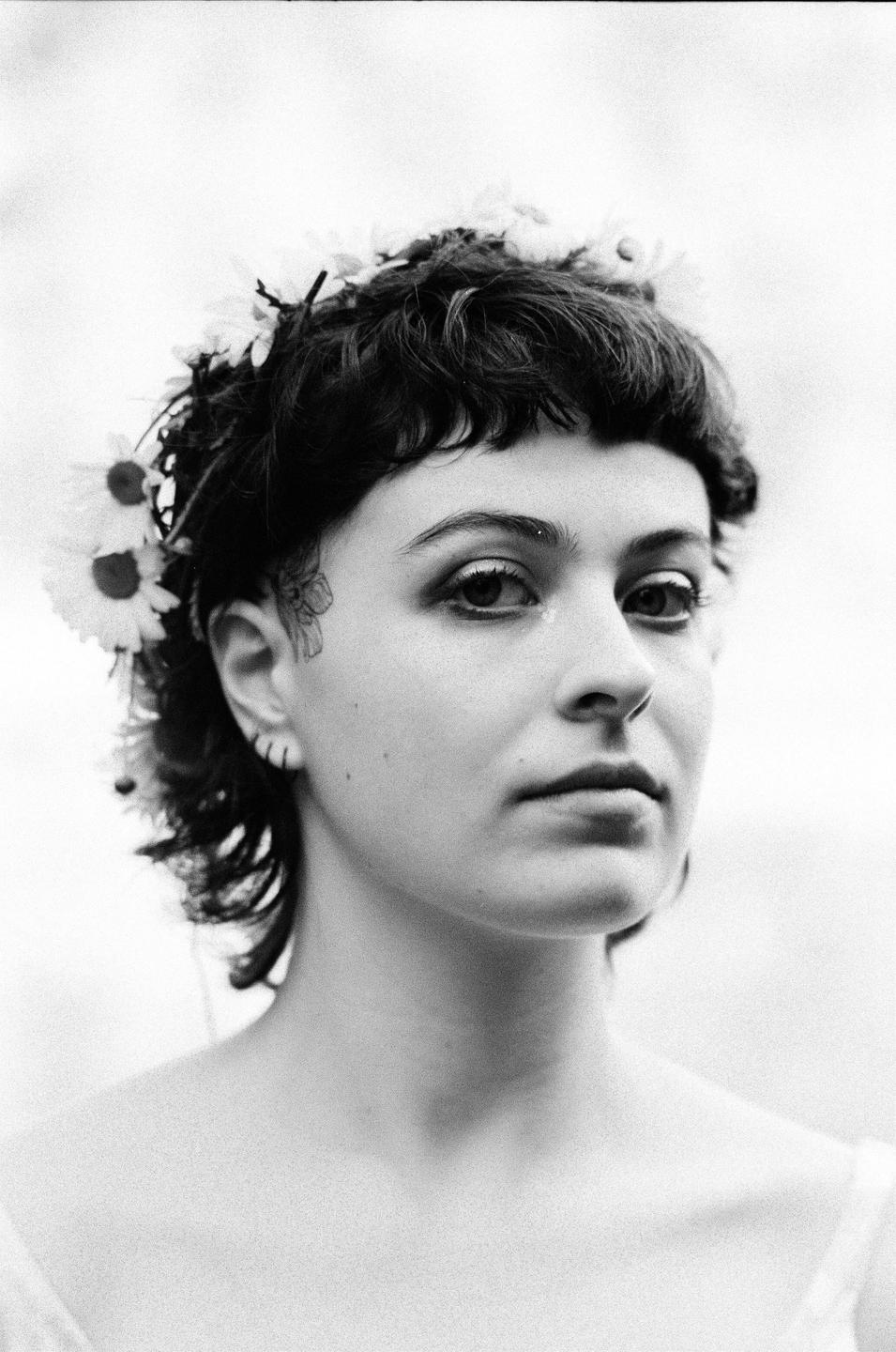 A woman with short curly hair and a flower crown looks directly at the viewer.