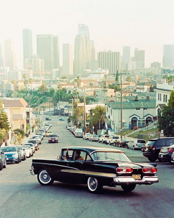 A black vintage car with tail fins drives up a hill in a city with a hazy skyline in the background.