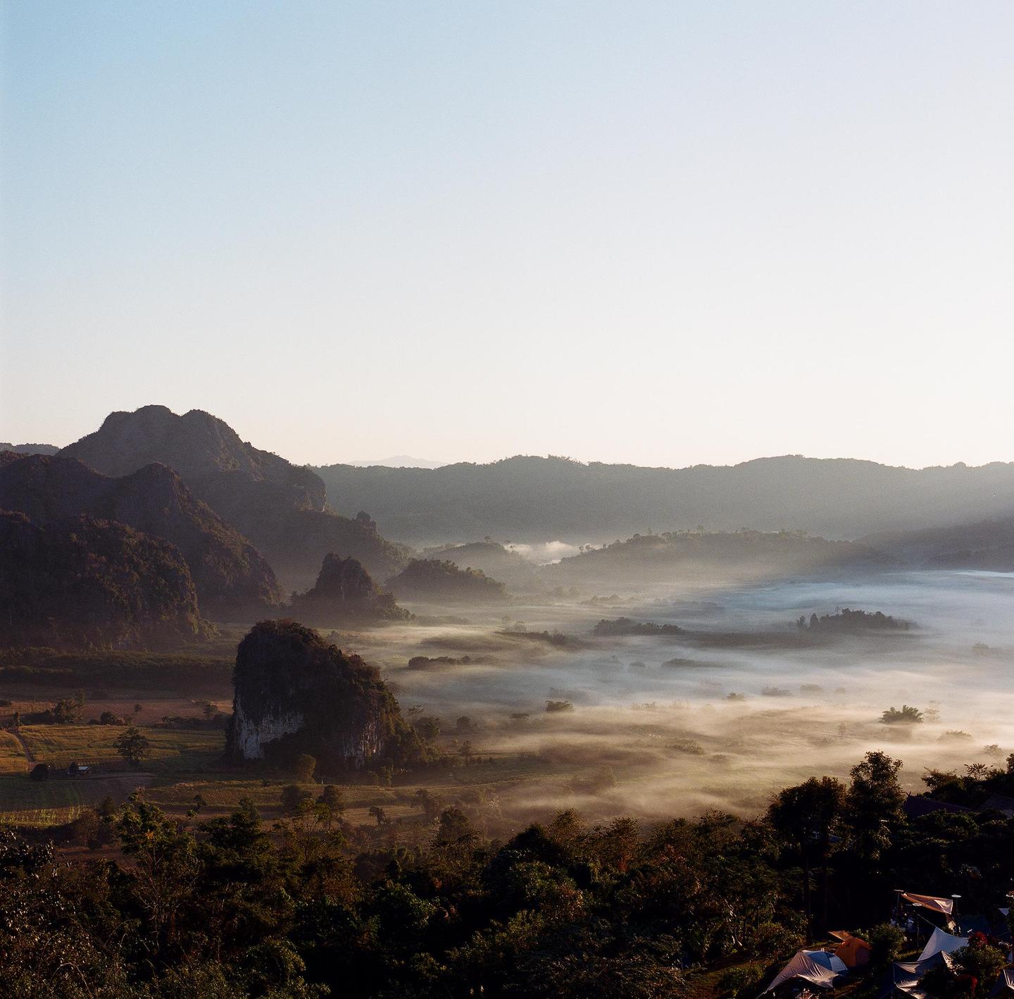 A wide landscape shows a valley filled with fog and surrounded by mountains under a clear sky.