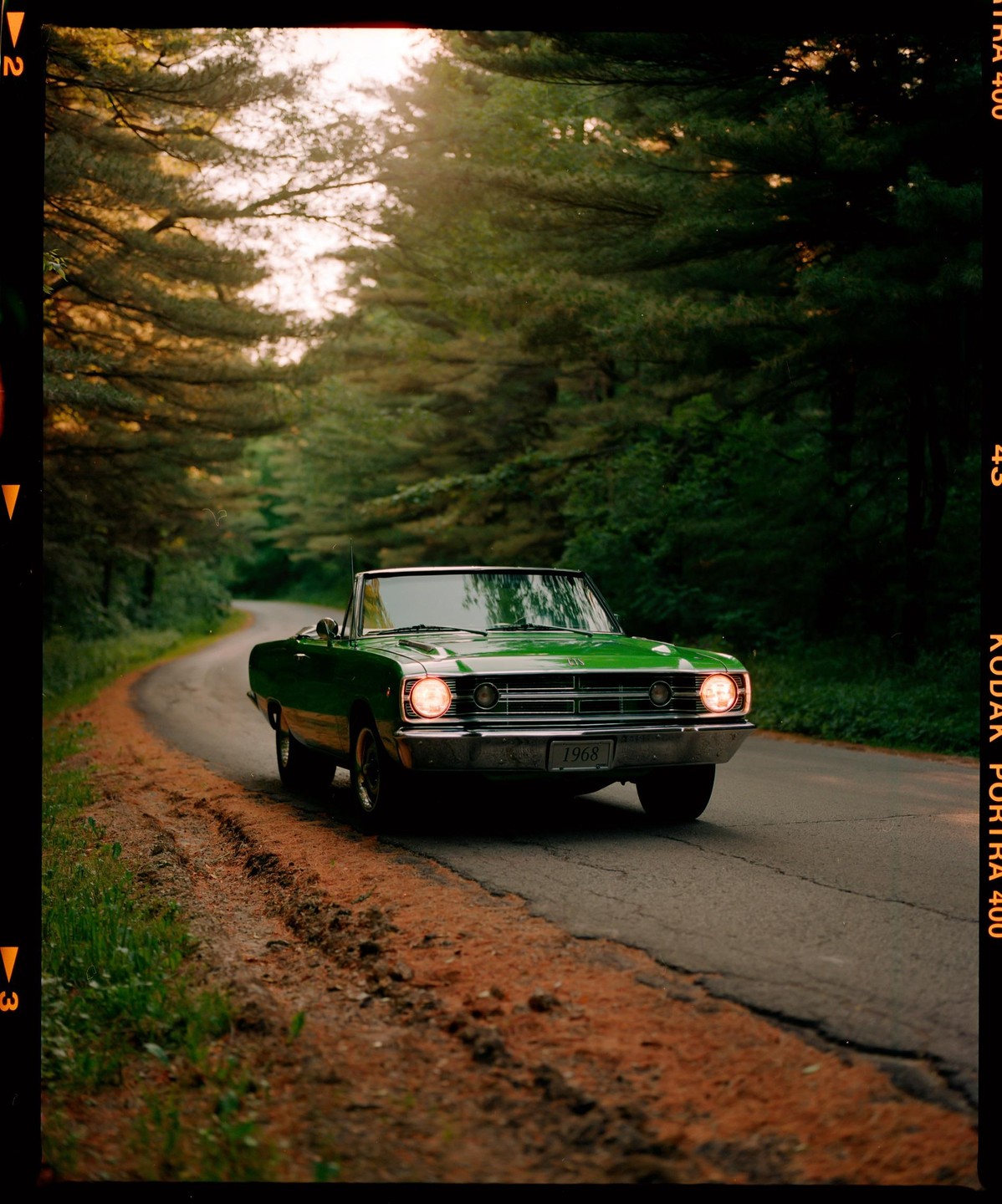 A green convertible car with its headlights on drives on a winding road through a forest.