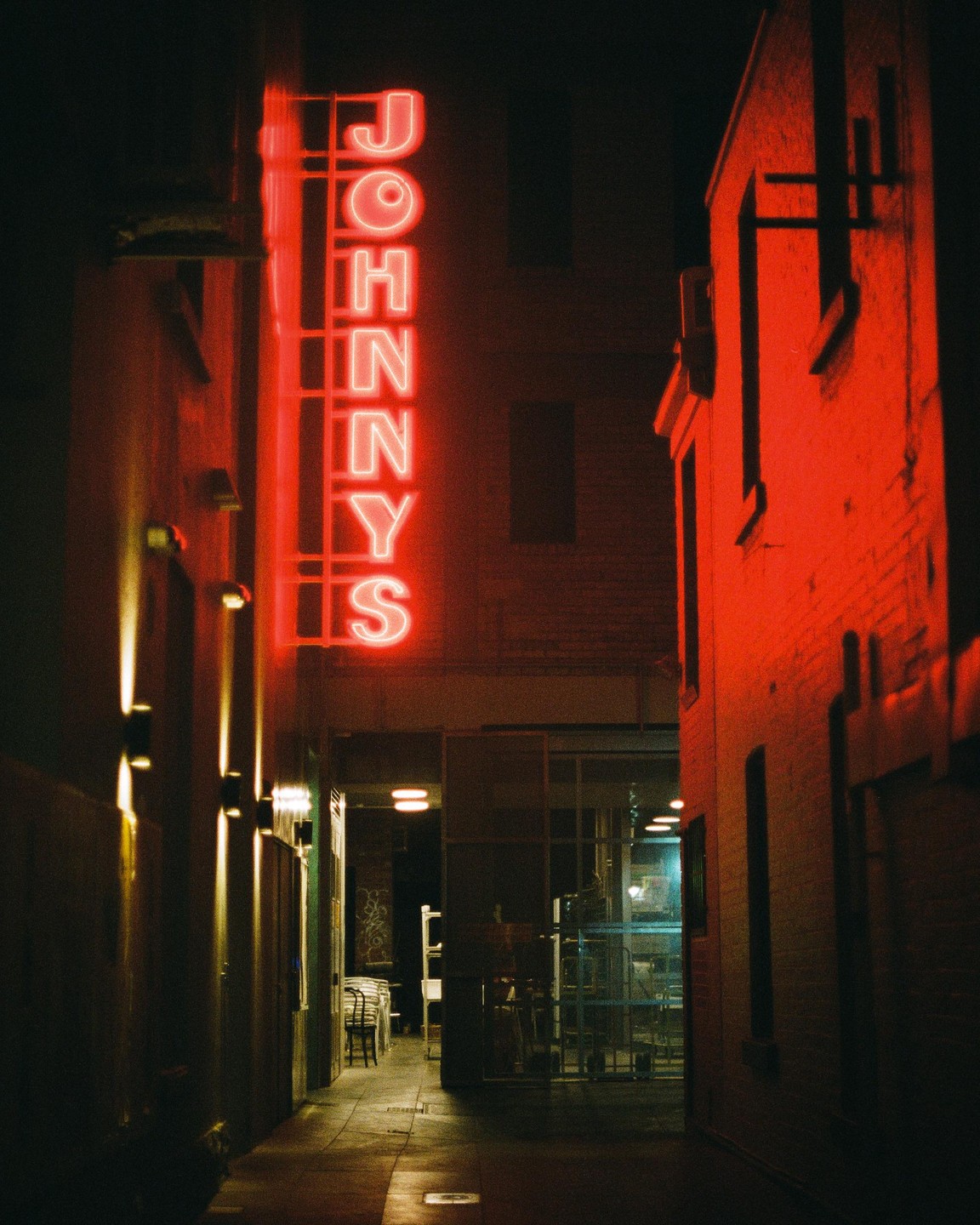 A red neon sign for "Johnny's" illuminates a narrow alleyway between two buildings at night.