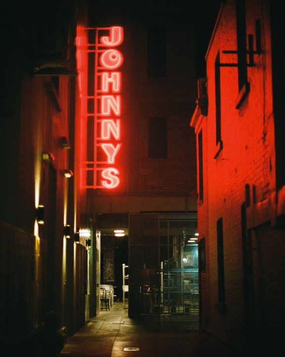 A red neon sign for "Johnny's" illuminates a narrow alleyway between two buildings at night.