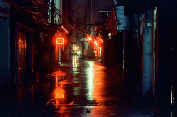 A narrow street is illuminated by neon signs and reflected in the wet pavement at night.