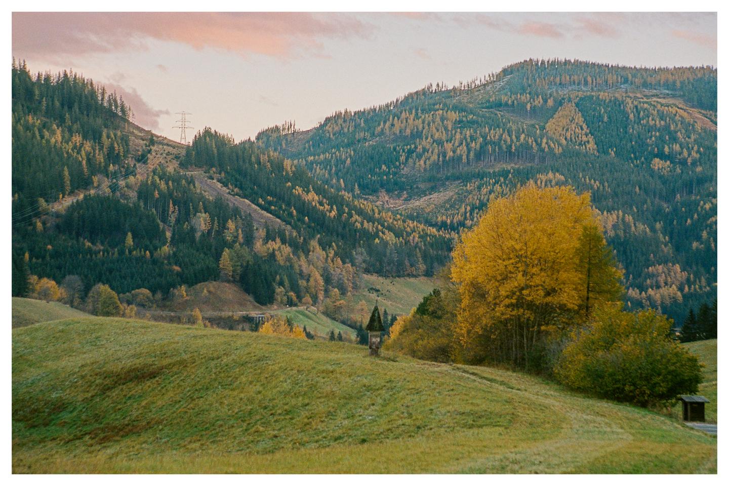 A small shrine sits on a grassy hill with a forest and mountains in the background under a pink sky.