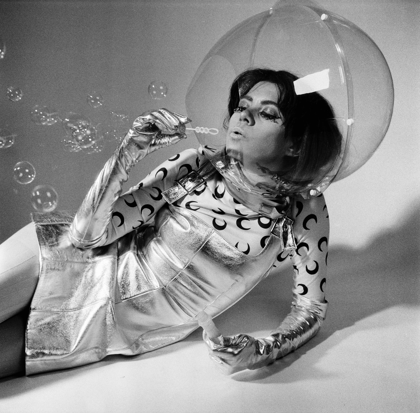 A woman in a futuristic outfit with a clear bubble helmet lies on the ground blowing bubbles.