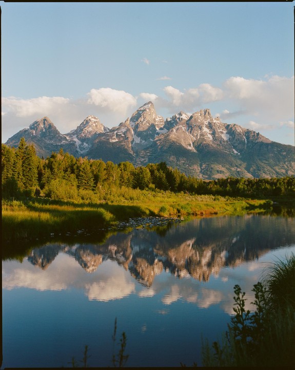 Snow-capped mountains are reflected in a calm body of water, with a line of green trees and grass in the middle ground under a blue sky with clouds.