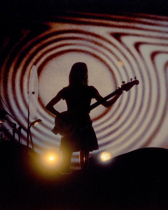 A woman in silhouette plays a bass guitar on a stage with a swirling pattern projected behind her.