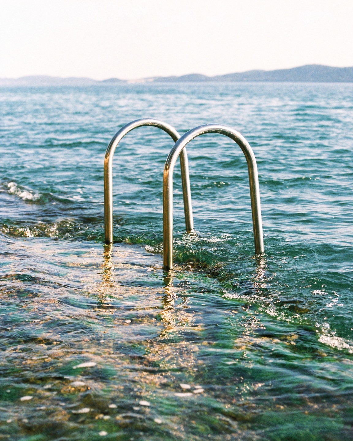 A metal ladder extends into the sea with mountains in the distance.