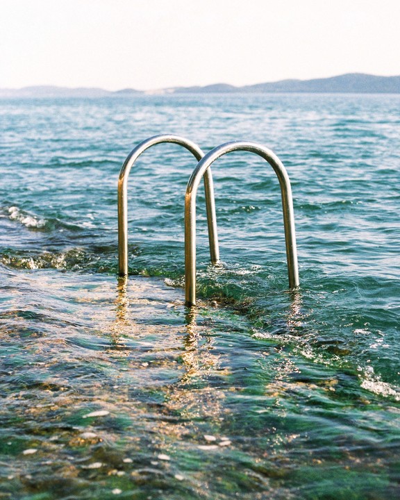 A metal ladder extends into the sea with mountains in the distance.