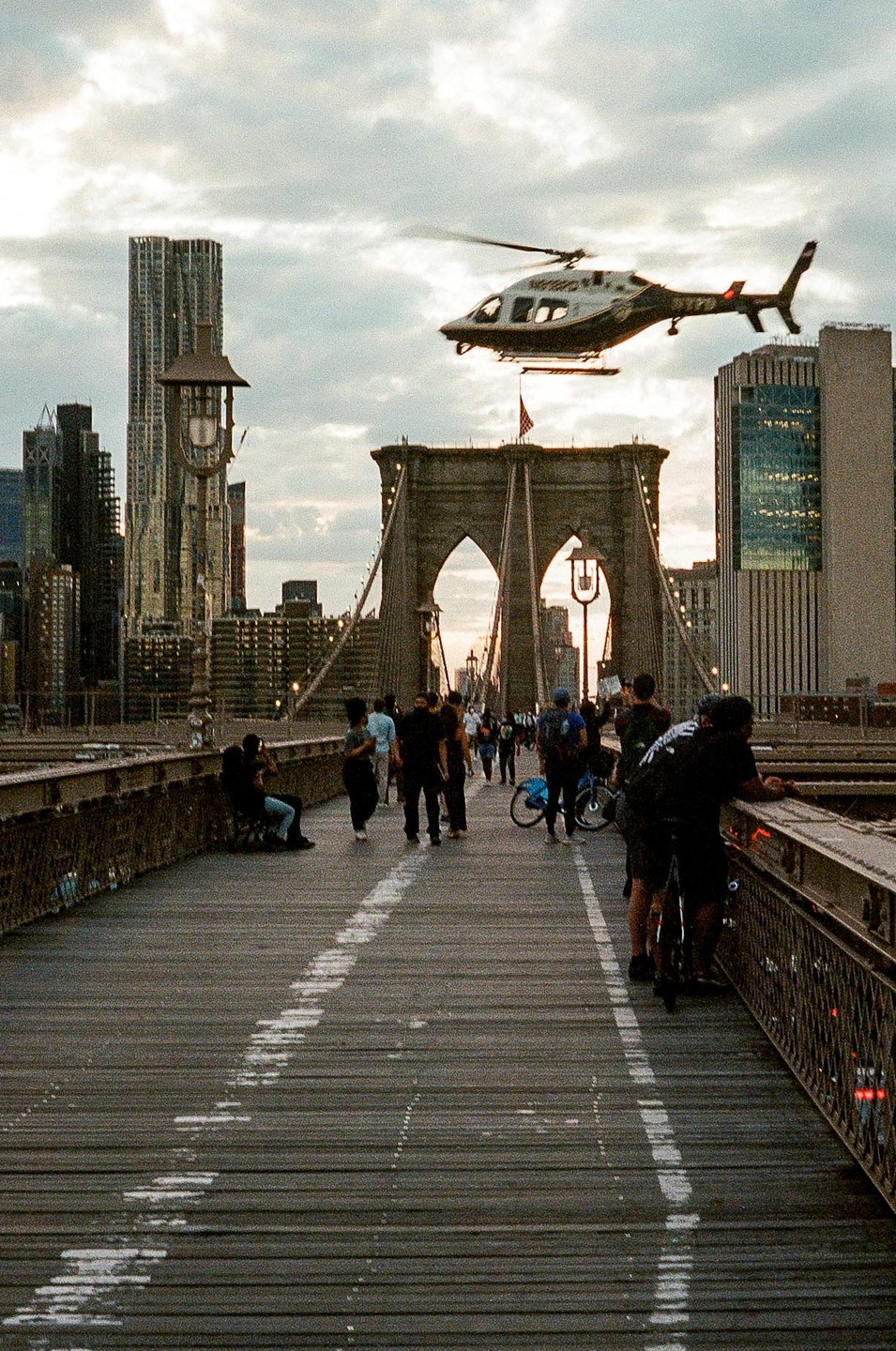 A helicopter flies over the Brooklyn Bridge, which is crowded with people, with the New York City skyline in the background.