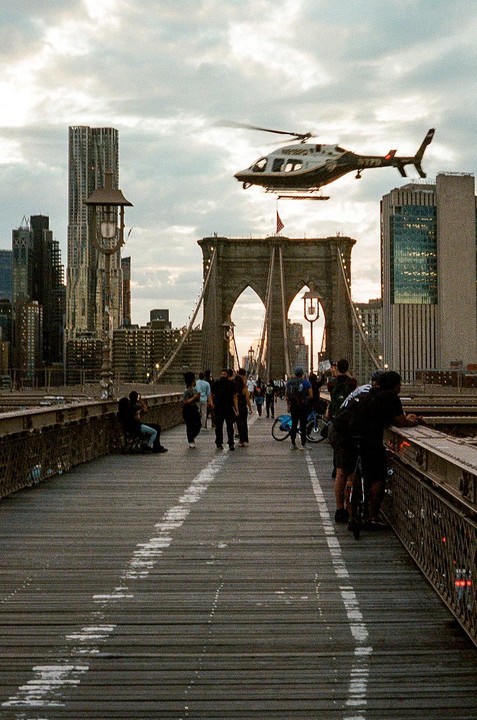 A helicopter flies over the Brooklyn Bridge, which is crowded with people, with the New York City skyline in the background.