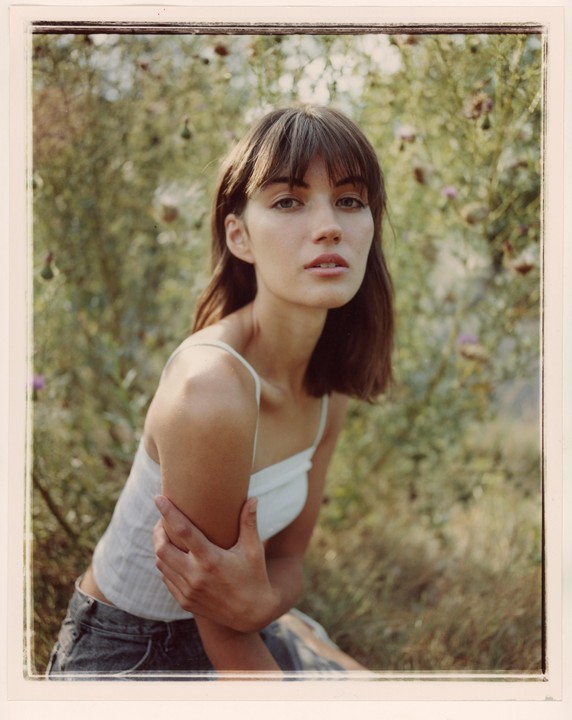 A woman with bangs and a white top is sitting in a field of green foliage with some purple flowers.