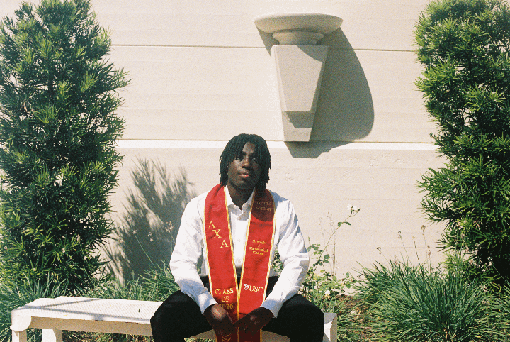 A man wearing a white shirt and a red graduation stole is sitting on a bench between two green bushes.