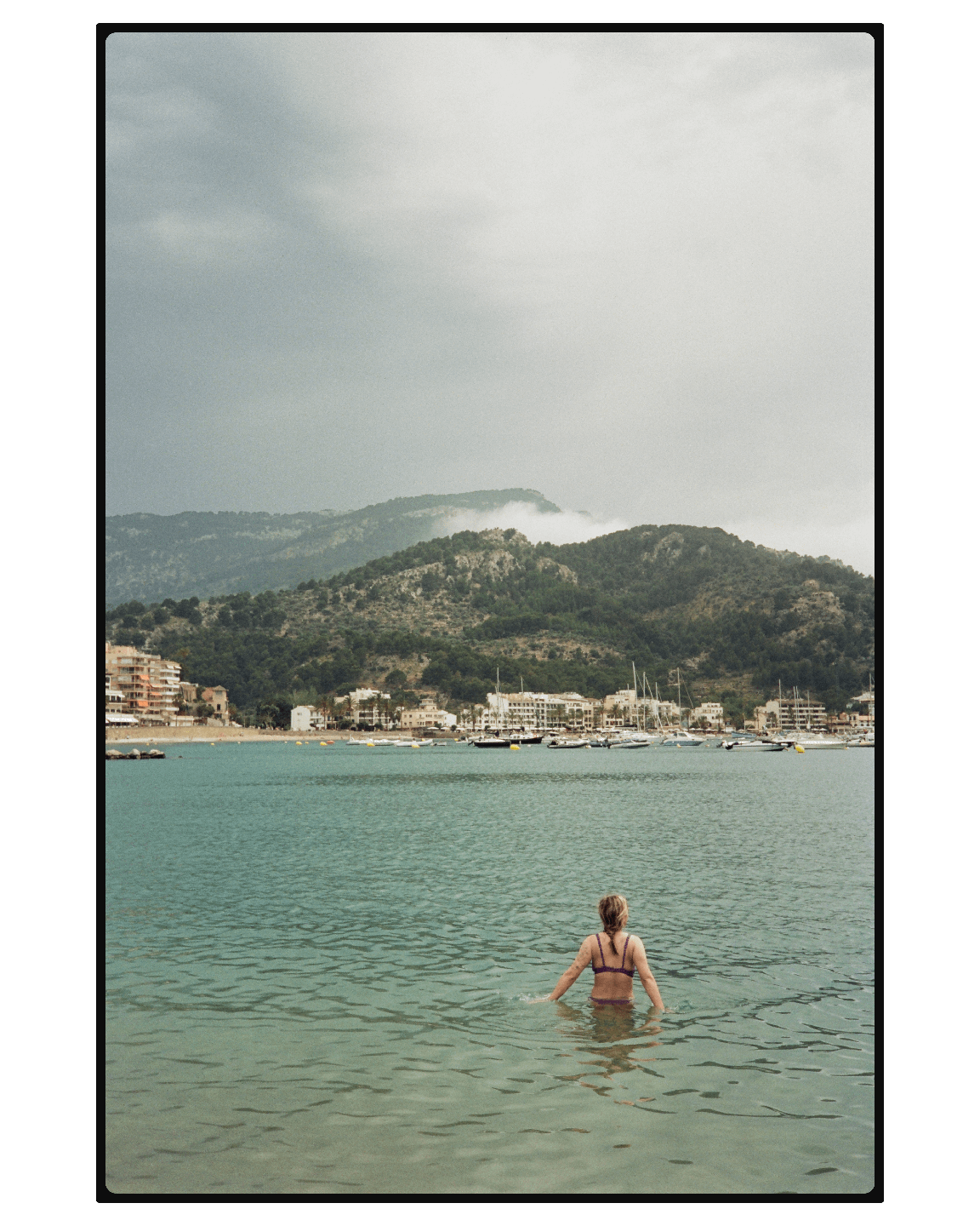A woman stands in the water of a bay with a town and mountains in the background.