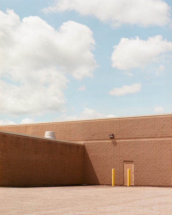 A building with textured walls and a light-colored door is visible under a blue sky with white clouds.