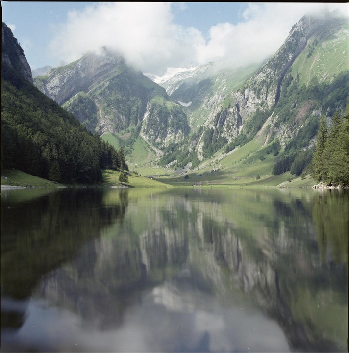 A calm lake reflects the surrounding mountains, some of which are topped with snow and partially obscured by clouds.