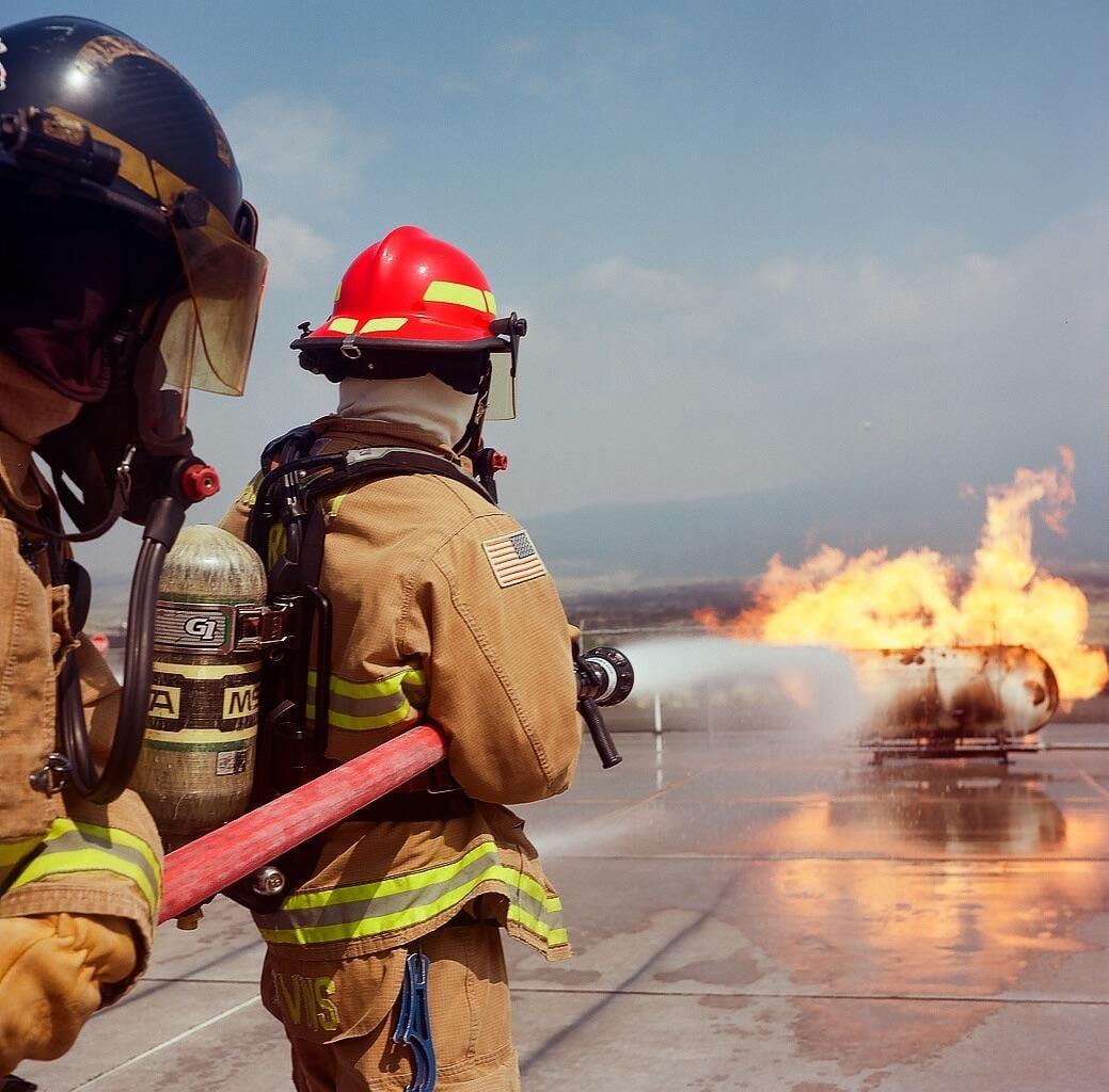 Two firefighters in full gear are extinguishing a large fire with a hose.