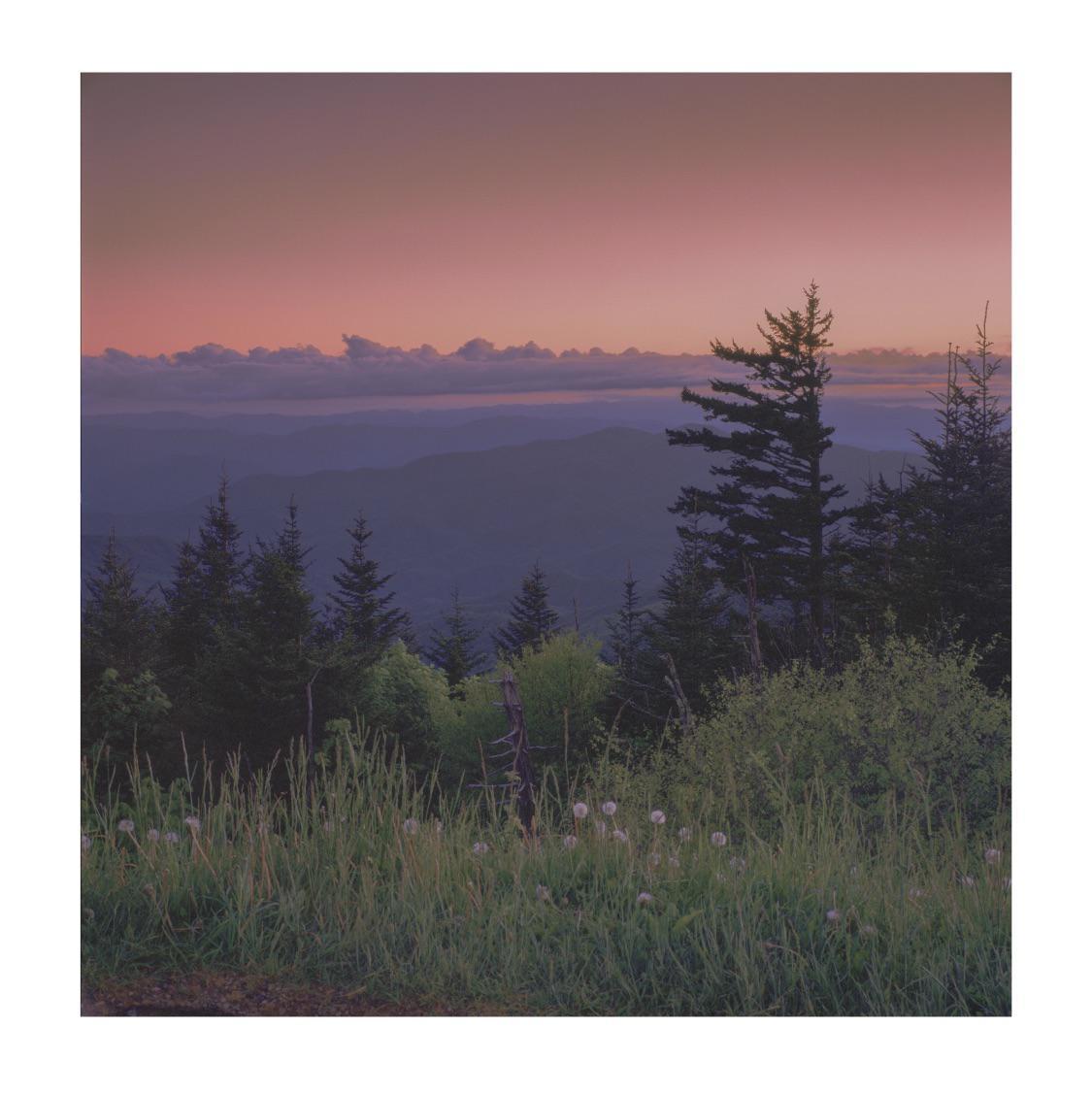 A mountain landscape at dusk shows a pink sky over layers of blue mountains and green trees.