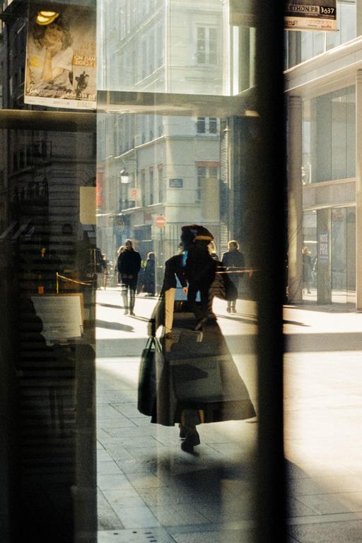 A woman walks down a sunlit street, reflected in a window.